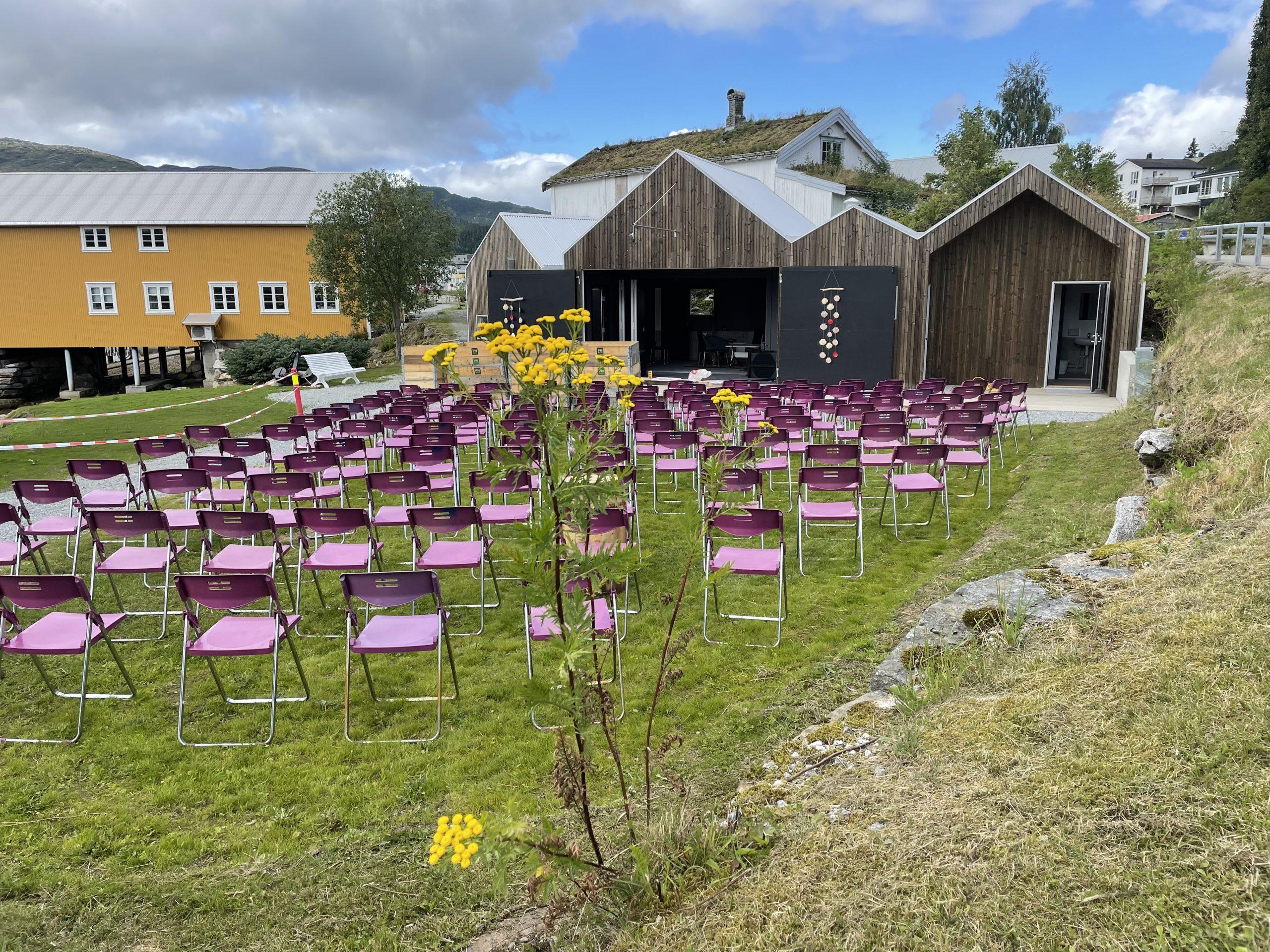 Utendørsscene med lilla stoler på gresset foran tre mørke trehus