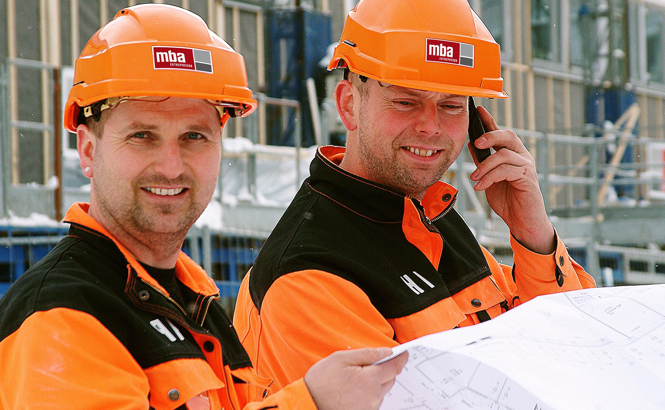 To smilende arbeidere i oransje jakker og MBA-hjelmer leser tegninger på byggeplassen
